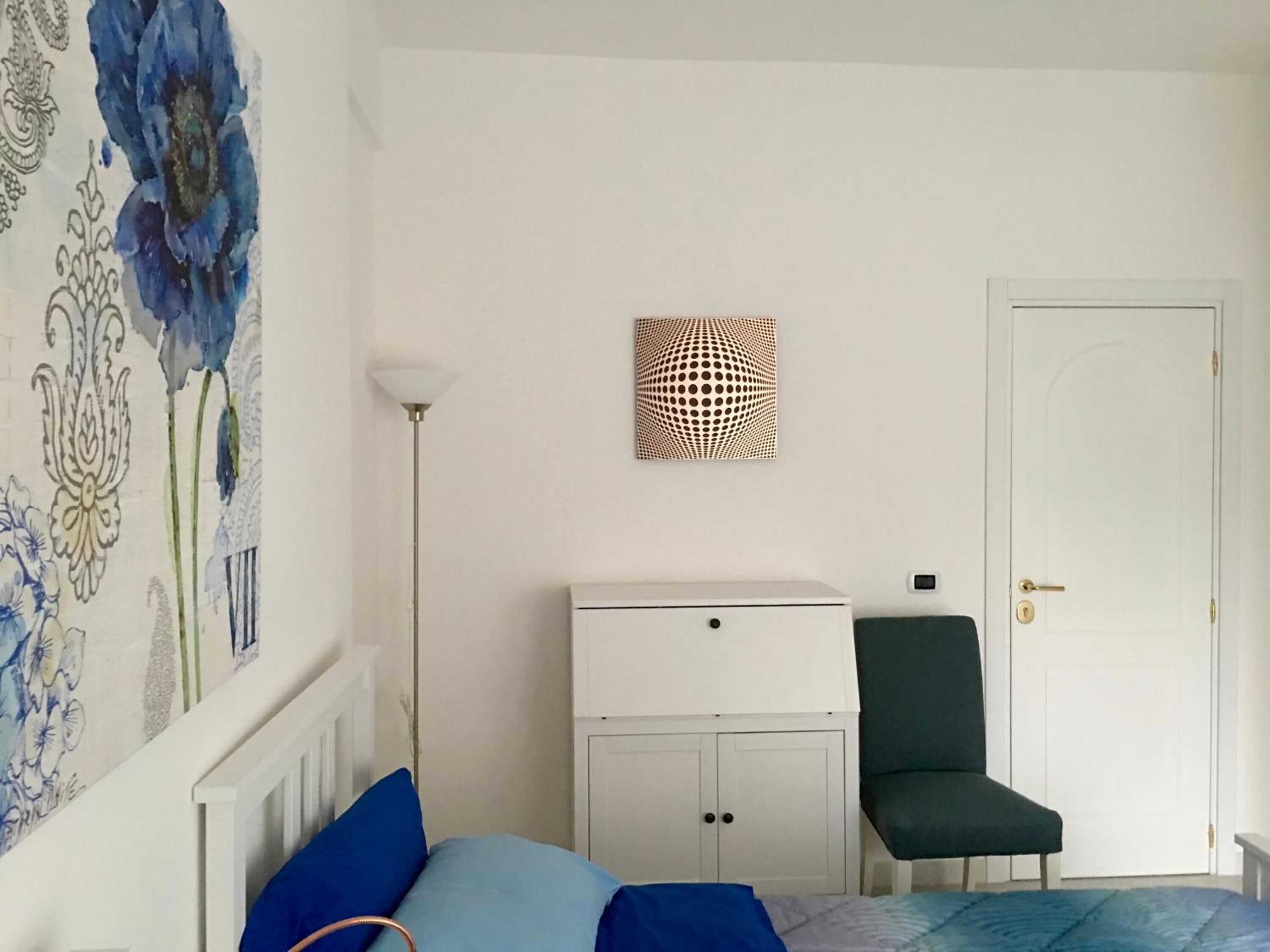A bedroom with a bed, a chair, a wardrobe, and a wall with a floral painting.
