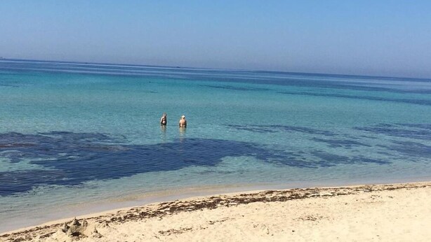 A beach with clear turquoise water and two people standing in the water. - Hibiscus (Taranto)