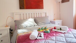 A neatly made bed with pillows, a bedside table with a lamp, and a tray with teapots and a rose. - Hibiscus (Taranto)