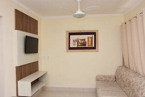 A living room with a flat-screen TV mounted on a wooden panel wall, a ceiling fan, a framed picture, and a patterned sofa. - Reserve Caldas Lacqua 1 (Caldas Novas)