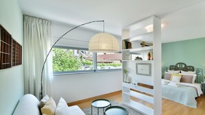 A modern bedroom with a large window, a white sofa, a bedside table, a small round table, and a bookshelf. - Villa Amaryllis (Athens)