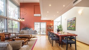 A spacious living area with a wooden floor, a dining table with chairs, a sofa set, and a large window providing natural light. - Villa Amaryllis (Athens)