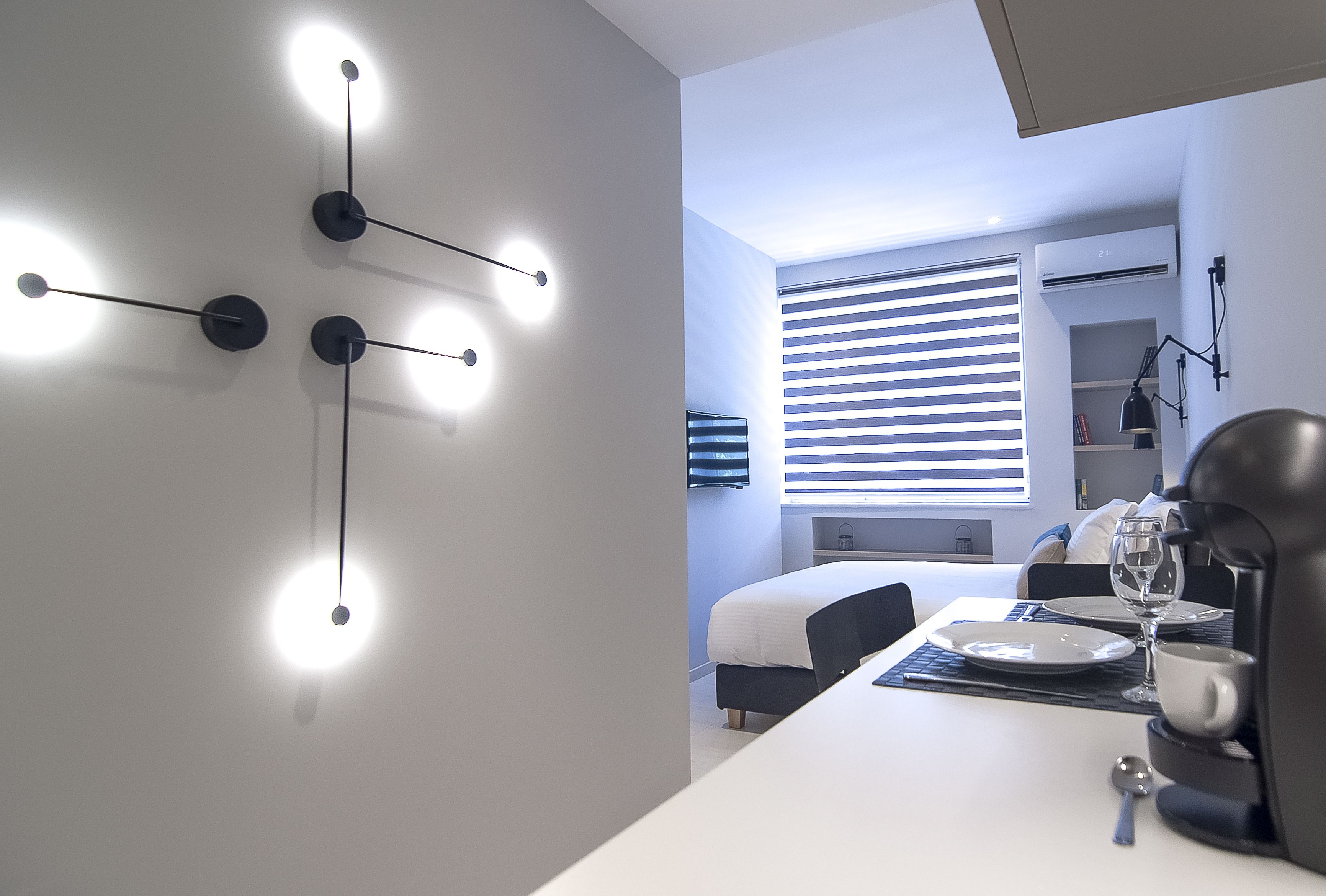 A modern hotel room with a white bed, a dining table set with plates and glasses, and a wall-mounted light fixture.
