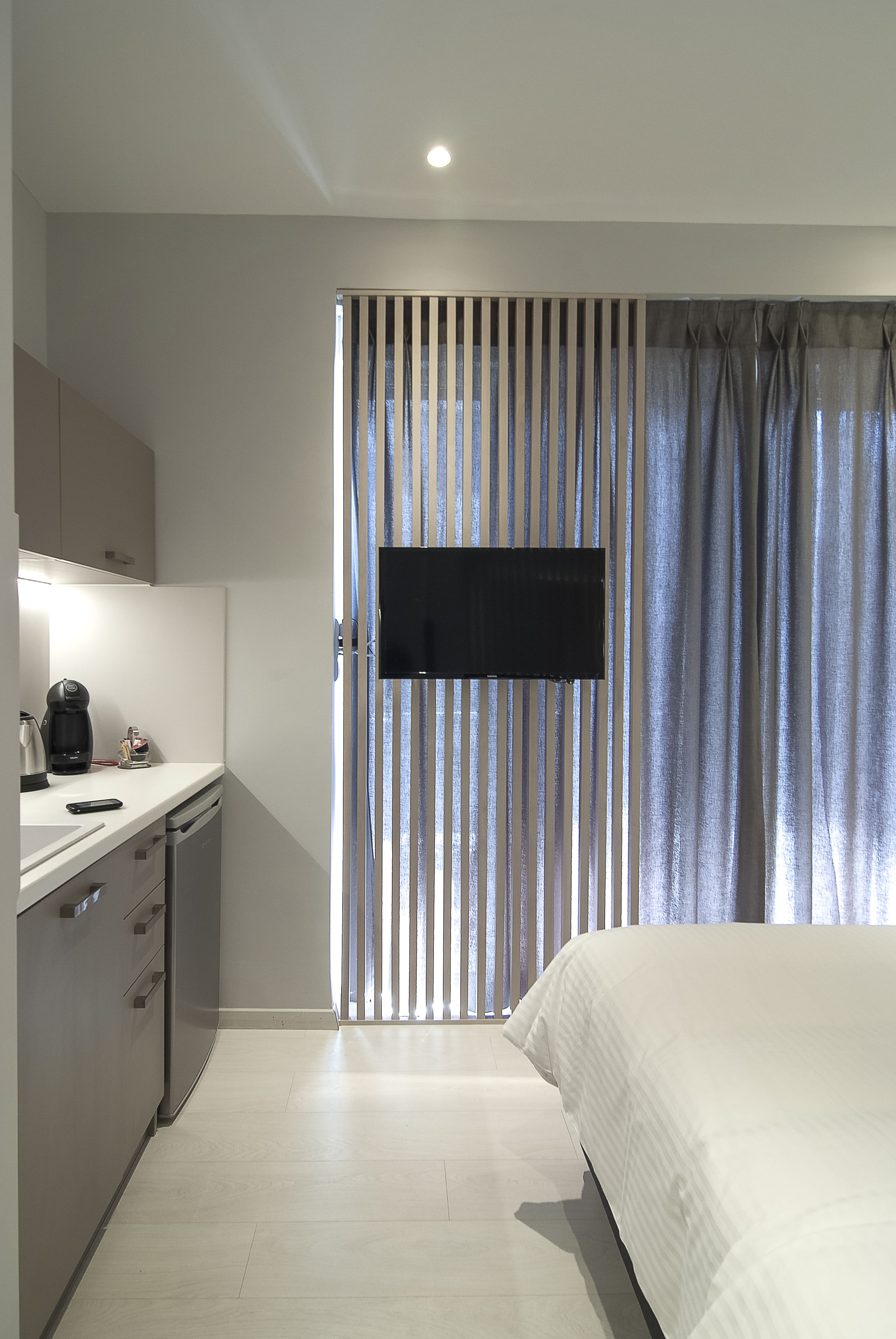 Quarto com cama, televisão na parede, cafeteira e área de cozinha com armários e bancada.