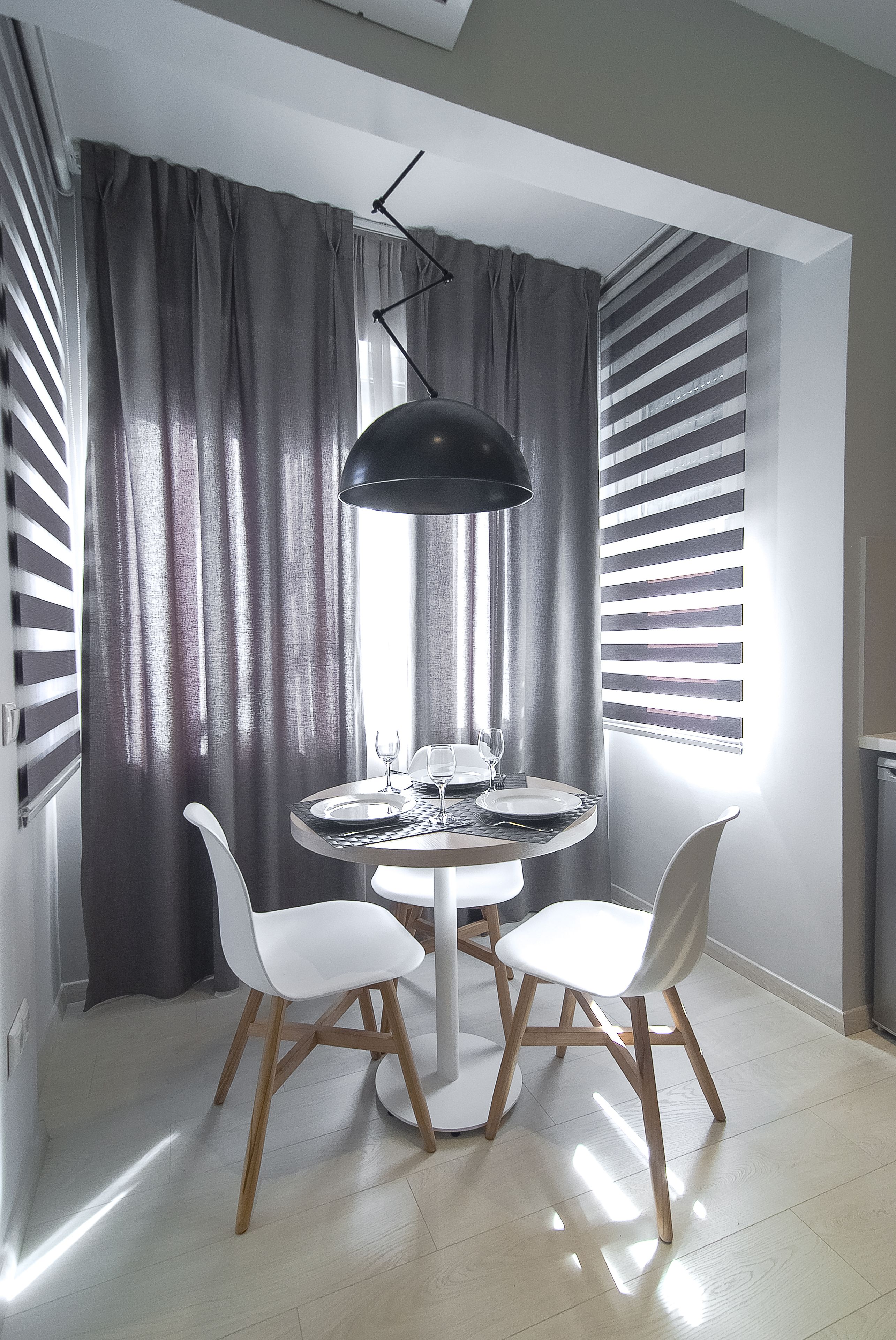 A dining area with a round table, two white chairs, a black pendant light, and a window with blinds.