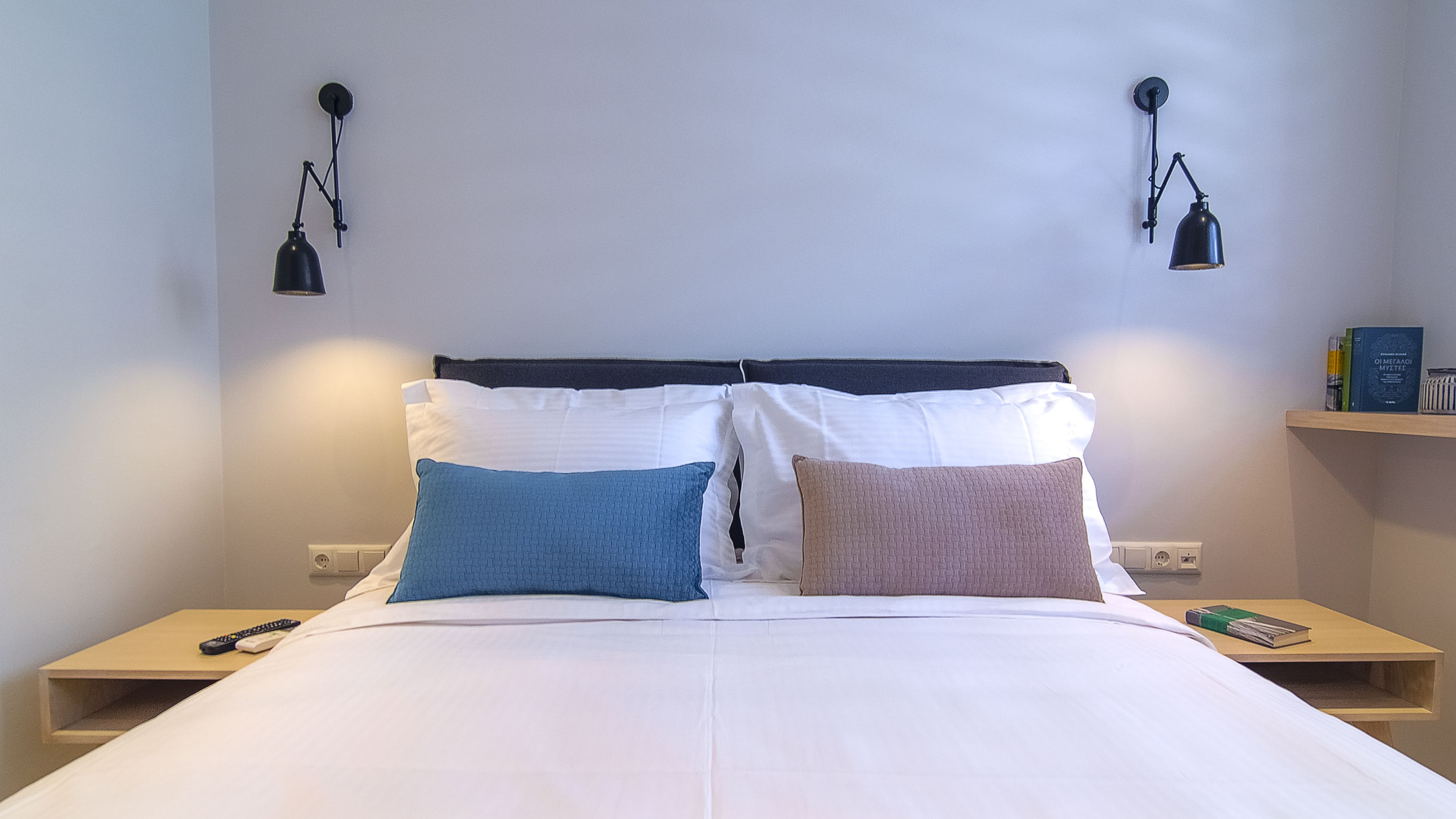 A neatly made bed with white bedding and two pillows, a bedside table with a book and a remote control, and two wall-mounted lamps.