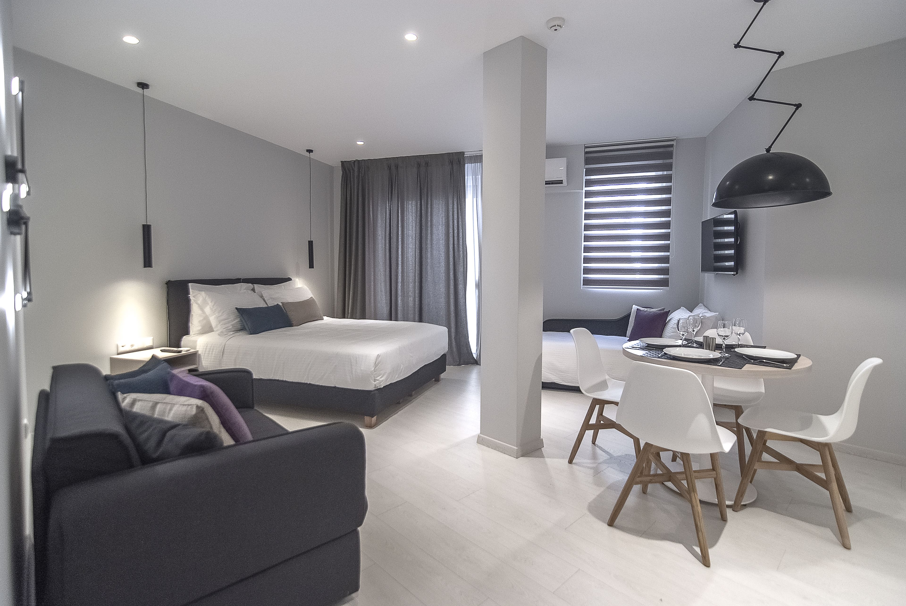 A modern hotel room with a bed, a sofa, a dining table with chairs, and a large window with blinds.