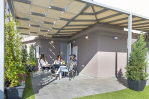Ein überdachter Patio-Bereich mit Tisch und Stühlen, umgeben von Topfpflanzen und einem gepflegten Rasen.