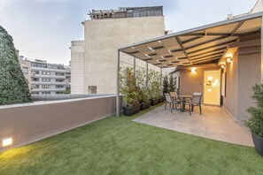 Eine Dachterrasse mit Kunstgrasanlage, Kübelpflanzen, einem Tisch und Stühlen sowie einer Pergola mit Beleuchtung.
