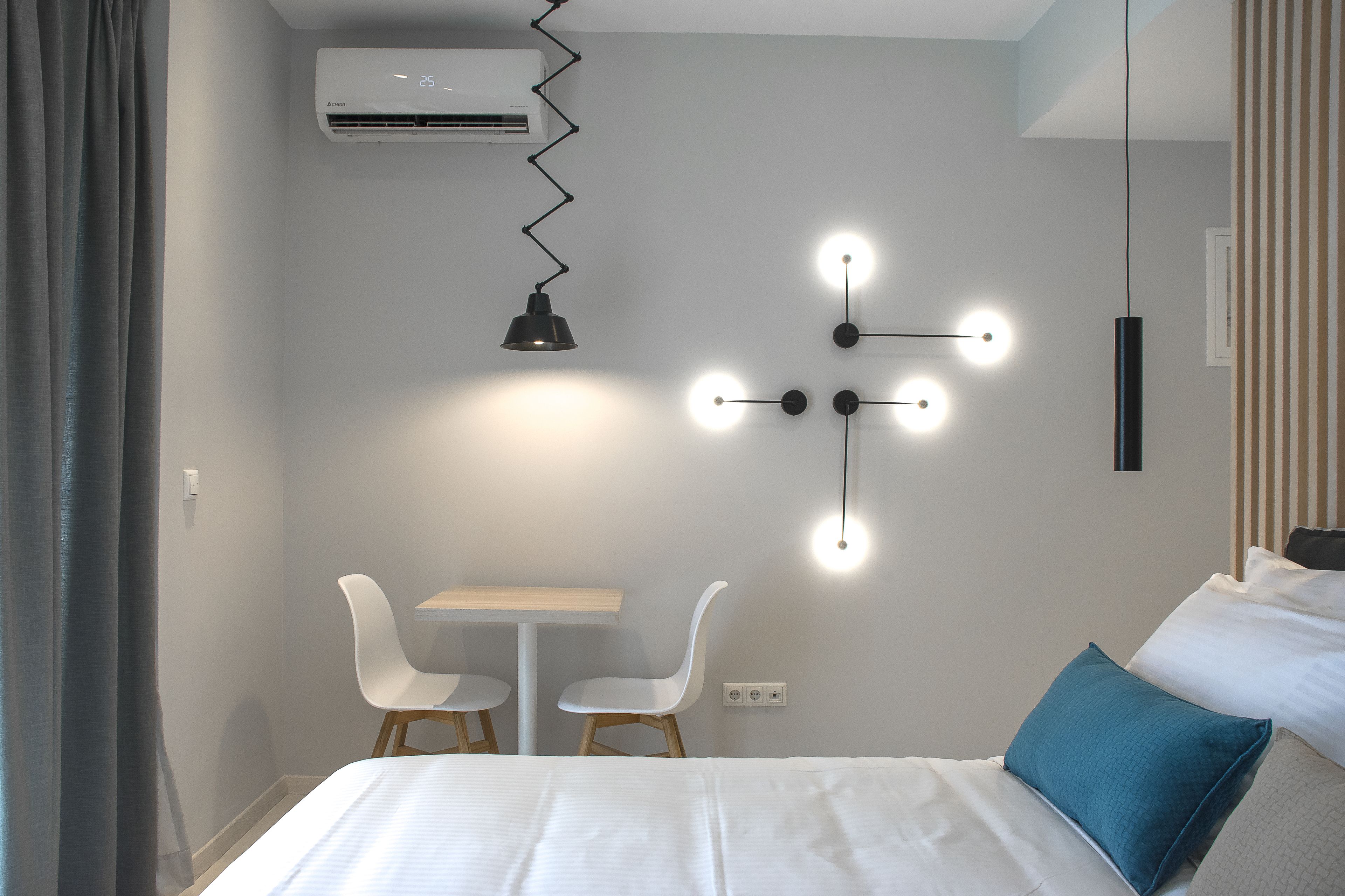A hotel room with a bed, a desk, two chairs, a wall-mounted air conditioner, and a unique lighting fixture.
