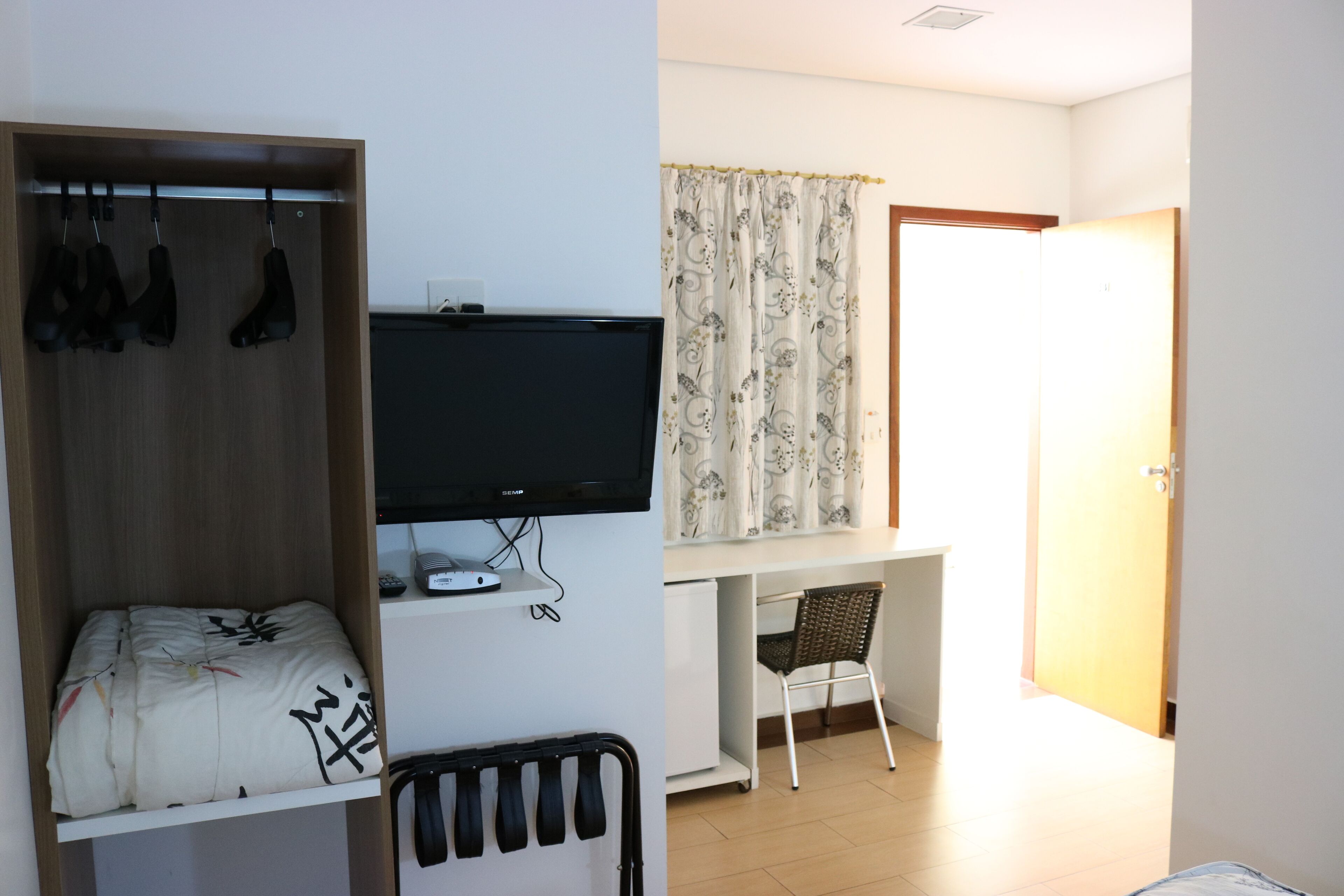 A room with a bed, a TV mounted on the wall, a desk with a chair, and a door leading to another room.