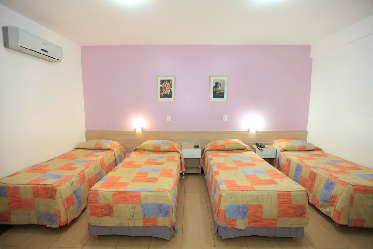 Une chambre d’hôtel avec quatre lits simples, chacun recouvert d’une couverture aux couleurs vives. Deux tableaux encadrés sont accrochés au mur, au-dessus des lits.