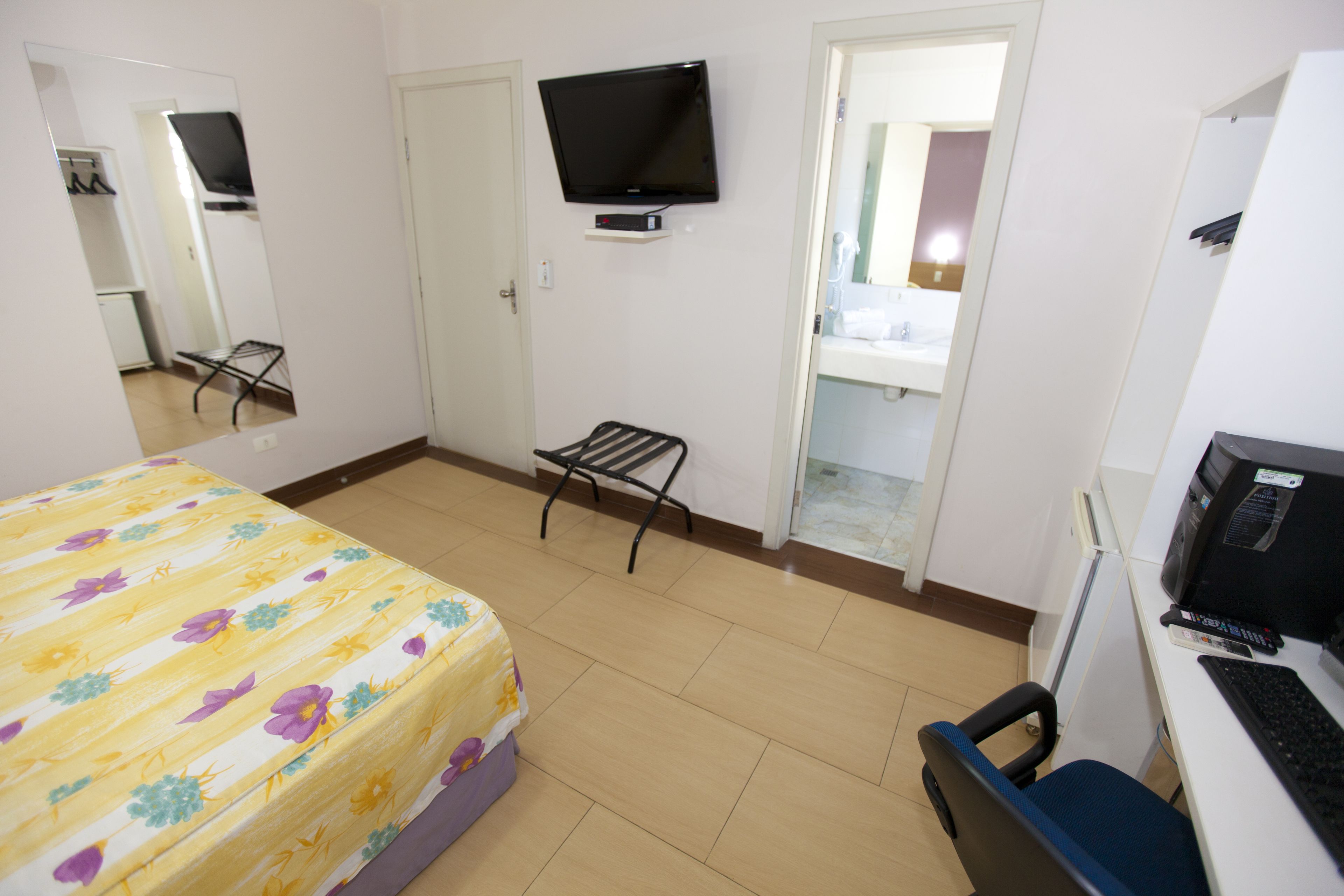 Quarto de hotel com cama, escrivaninha com computador, televisão fixada na parede e porta para o banheiro.