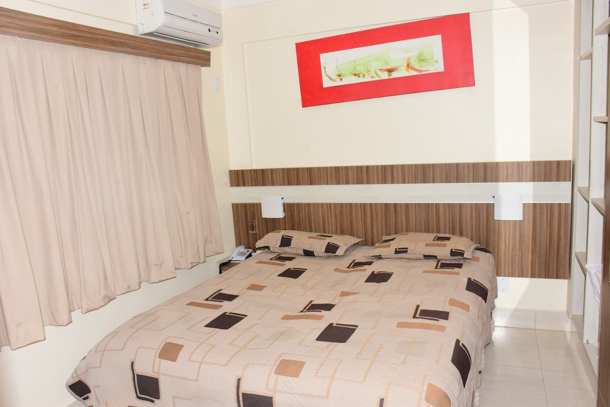 A bedroom with a bed, curtains, a wall-mounted air conditioner, and a framed picture.