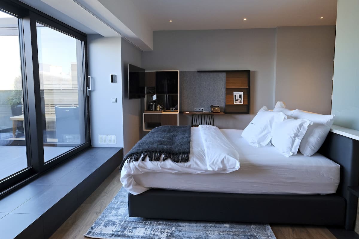 A modern bedroom with a large bed, a desk, and a view through a sliding glass door.