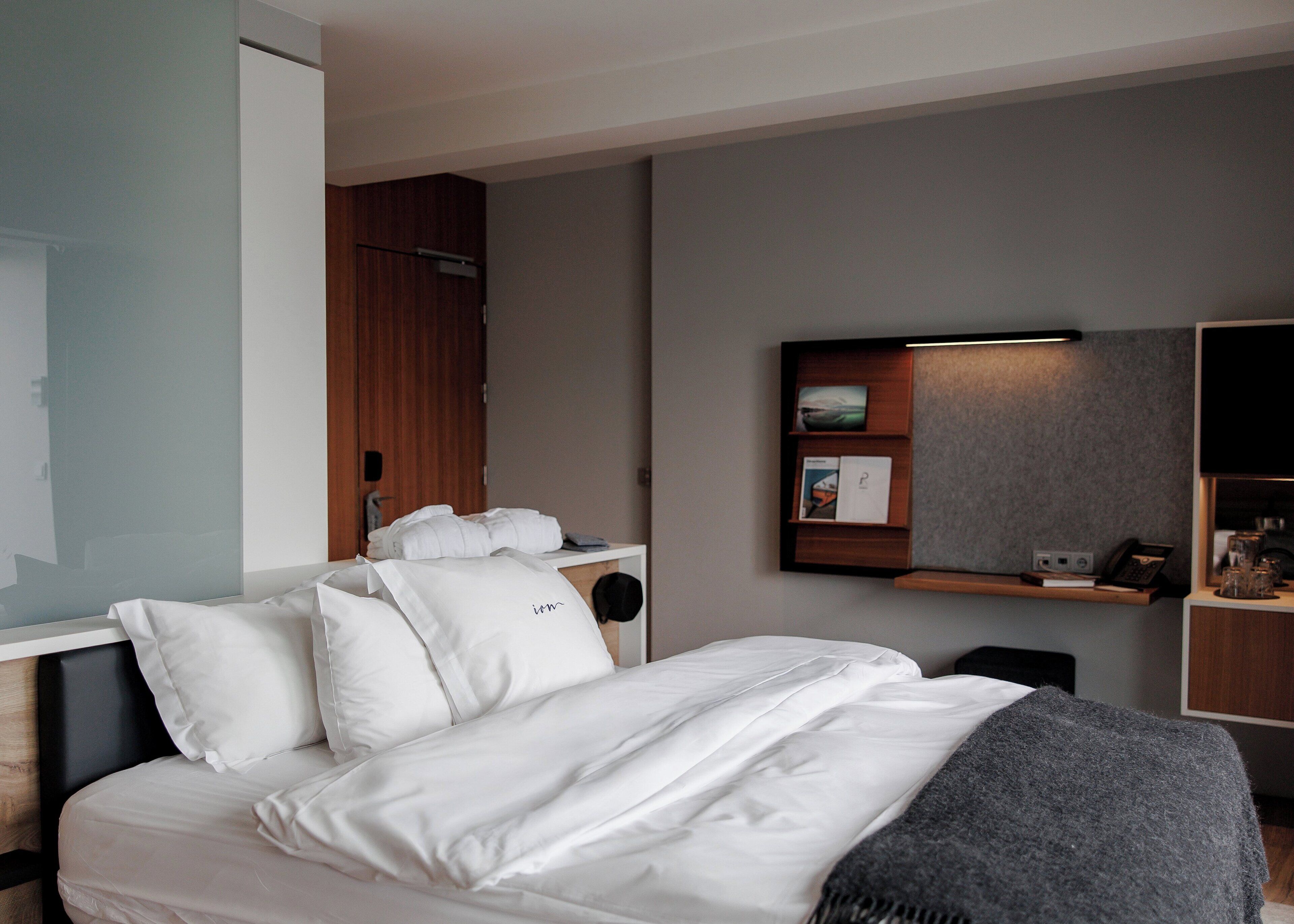 A modern hotel room with a neatly made bed, a bedside table with a lamp, a small shelf with books and a phone, and a wall-mounted TV.