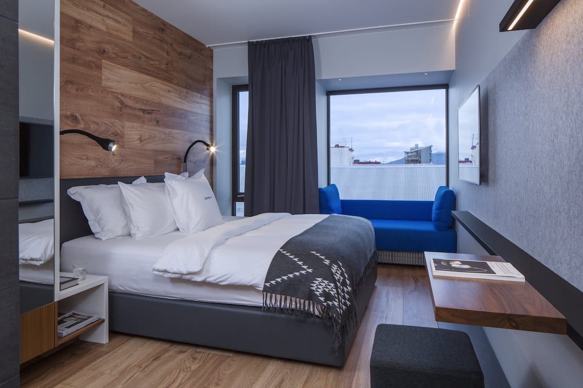 A modern hotel room with a large bed, a blue sofa, a wooden accent wall, and a view of the city.