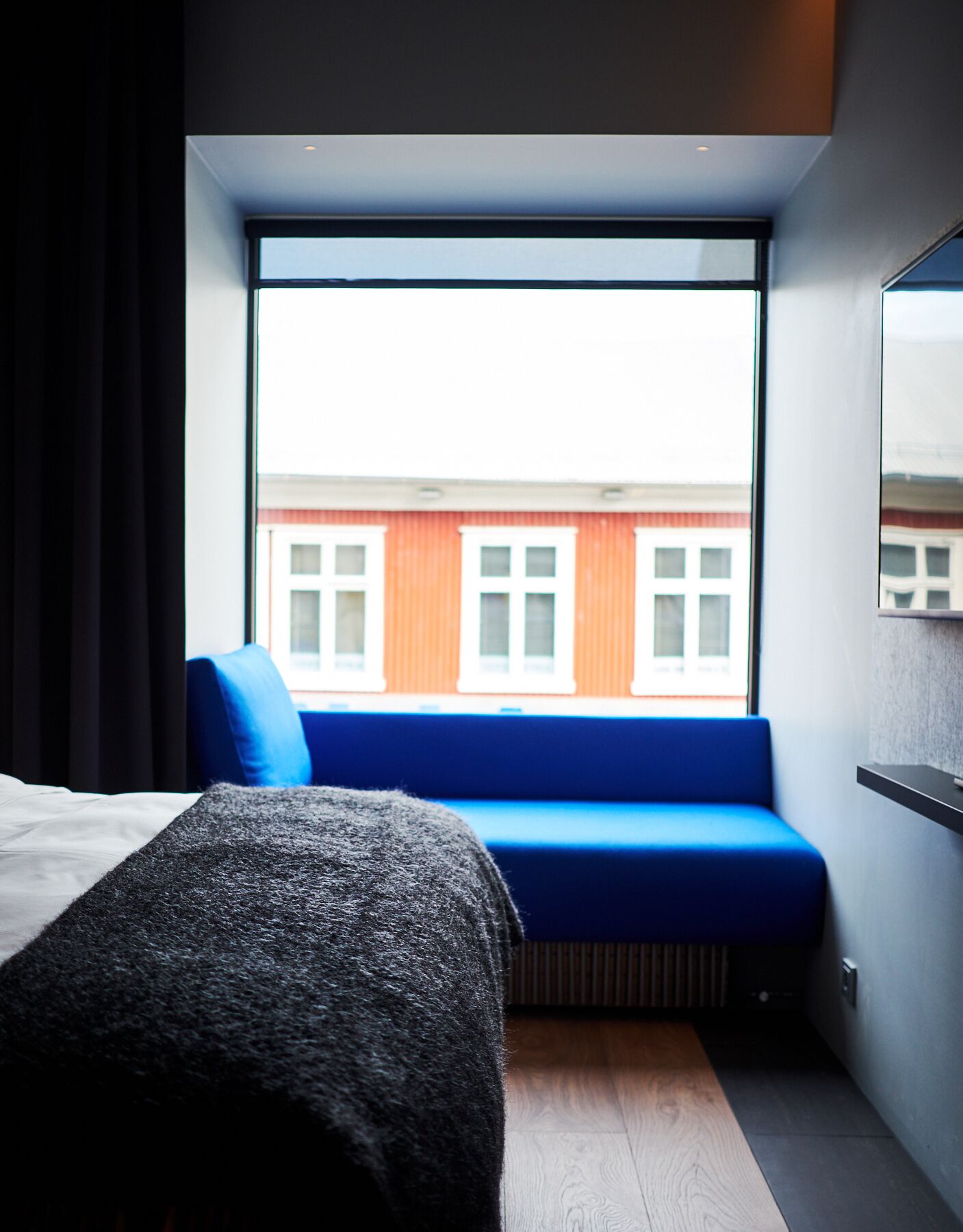 A modern hotel room with a large window, a blue sofa, a bed, and a wooden floor.