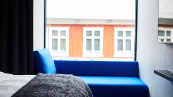 A modern hotel room with a large window, a blue sofa, a bed, and a wooden floor.