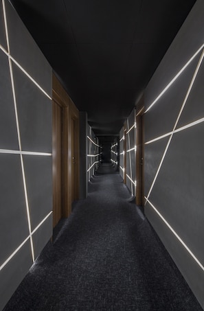 Un pasillo con alfombra oscura, puertas de madera y paredes con patrones geométricos iluminados.