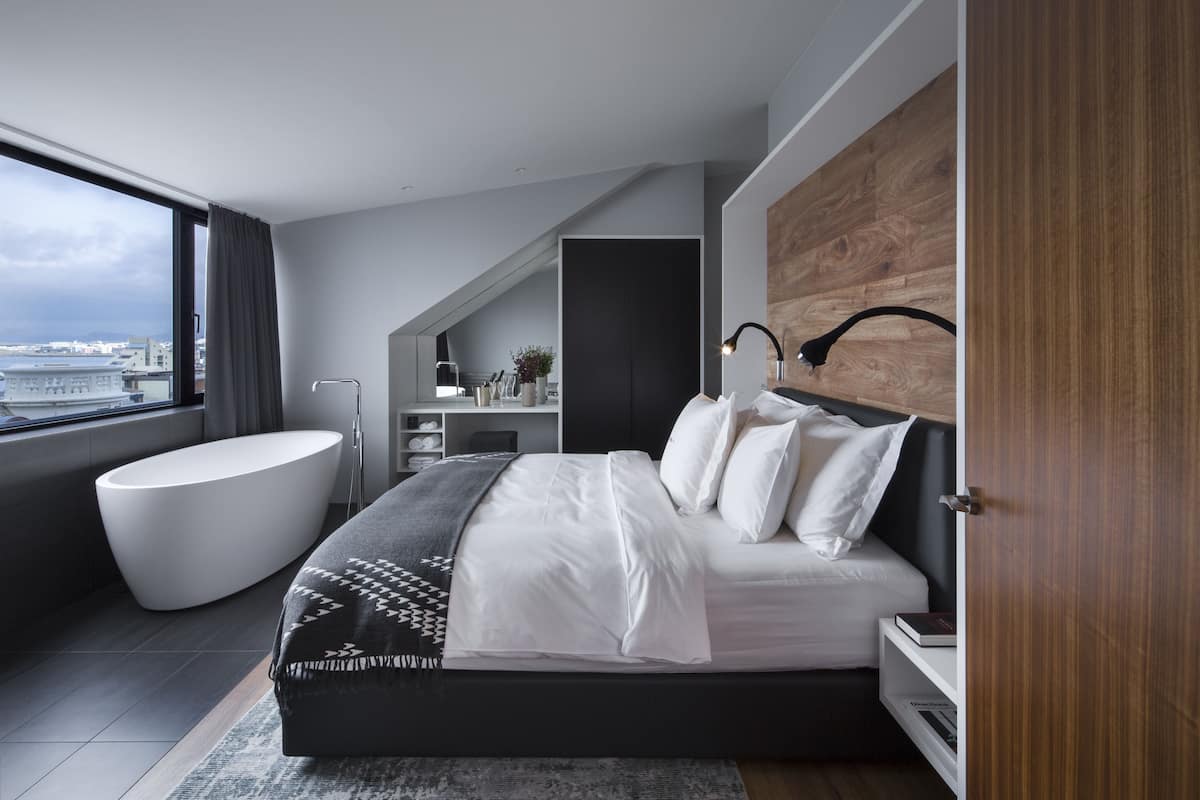 A modern bedroom with a large bed, a built-in bench, a bathtub, and a view of the city.