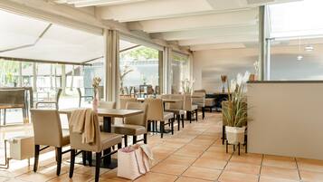 A spacious indoor area with multiple tables and chairs, large windows, and potted plants.