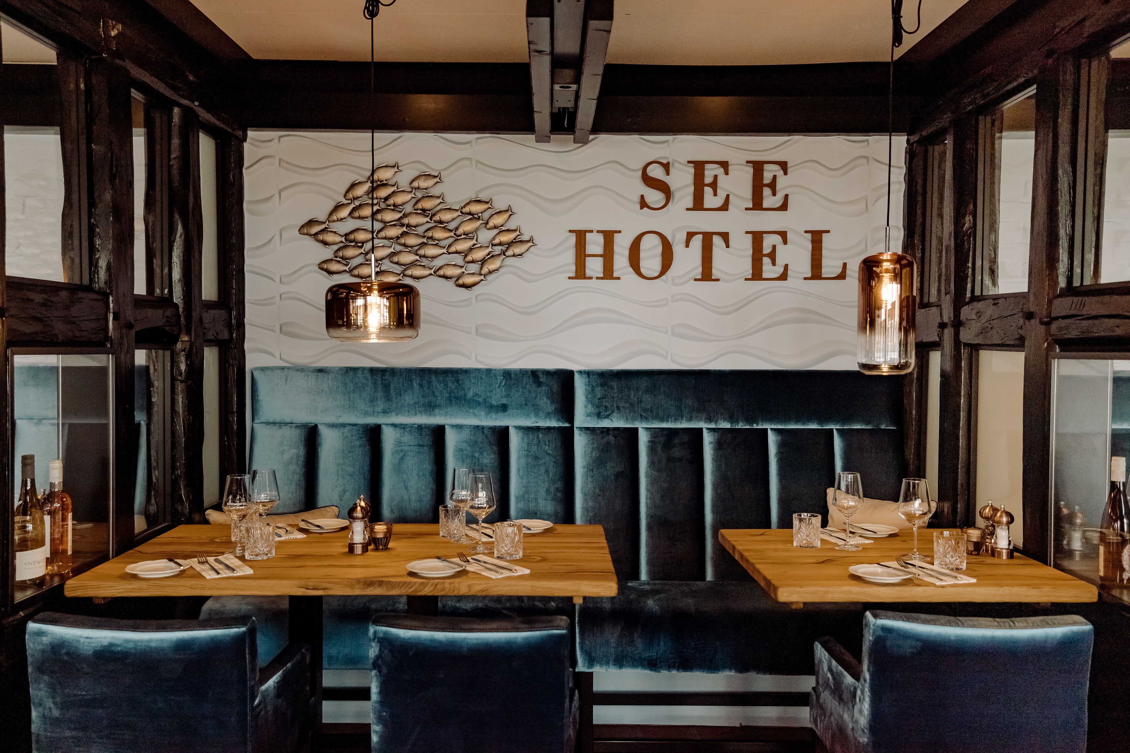 Um restaurante com mesa de madeira preparada para dois, um assento estofado azul e uma parede com o texto "SEE HOTEL".