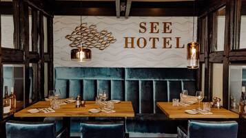 Um restaurante com mesa de madeira preparada para dois, um assento estofado azul e uma parede com o texto "SEE HOTEL".