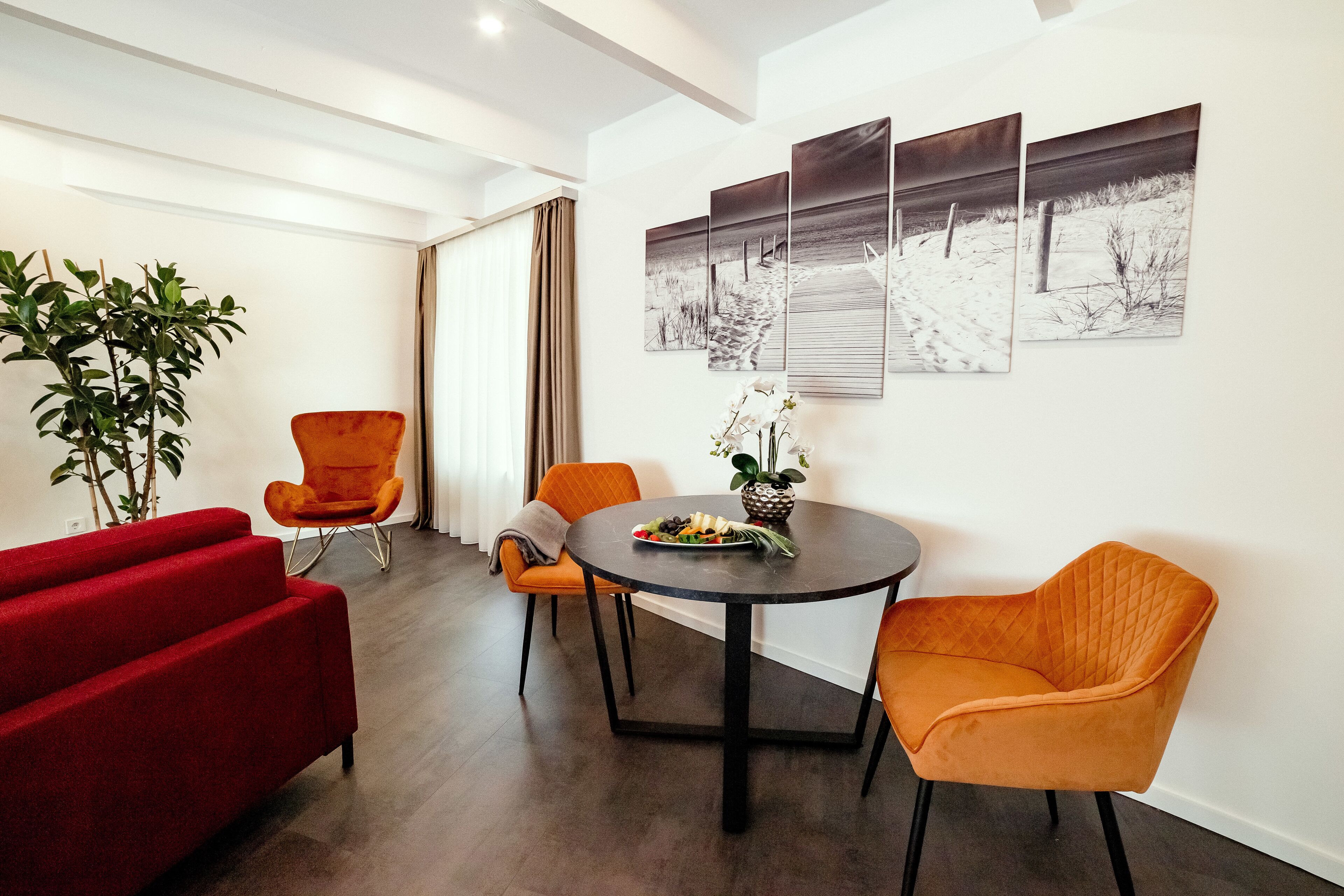 Una sala de estar moderna con un sofá rojo, dos sillones naranjas, una mesa redonda con un jarrón de flores y un ventanal con cortinas.