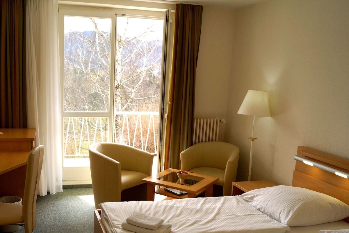 Habitación de hotel con cama, dos sillas, escritorio, lámpara y vista a árboles por la ventana.