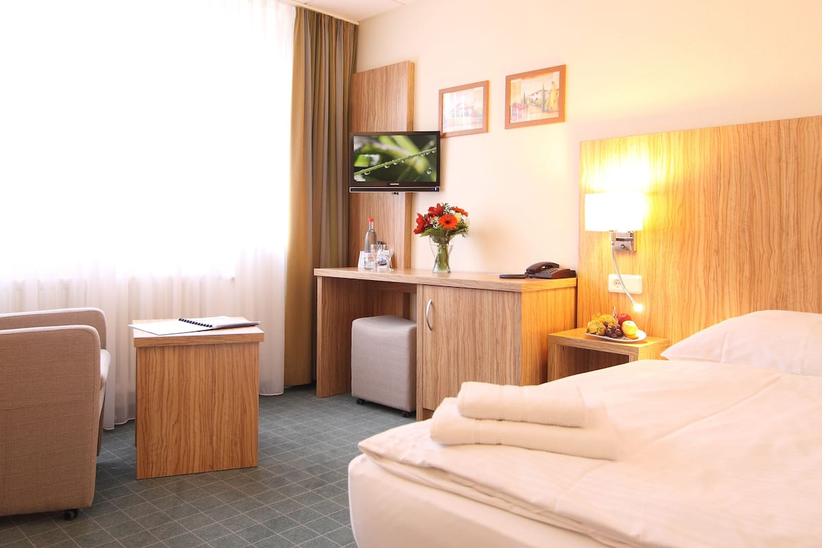 Une chambre d’hôtel comprenant un lit, un bureau avec une chaise, une télévision, une petite table avec un vase de fleurs et une table de chevet avec une lampe.