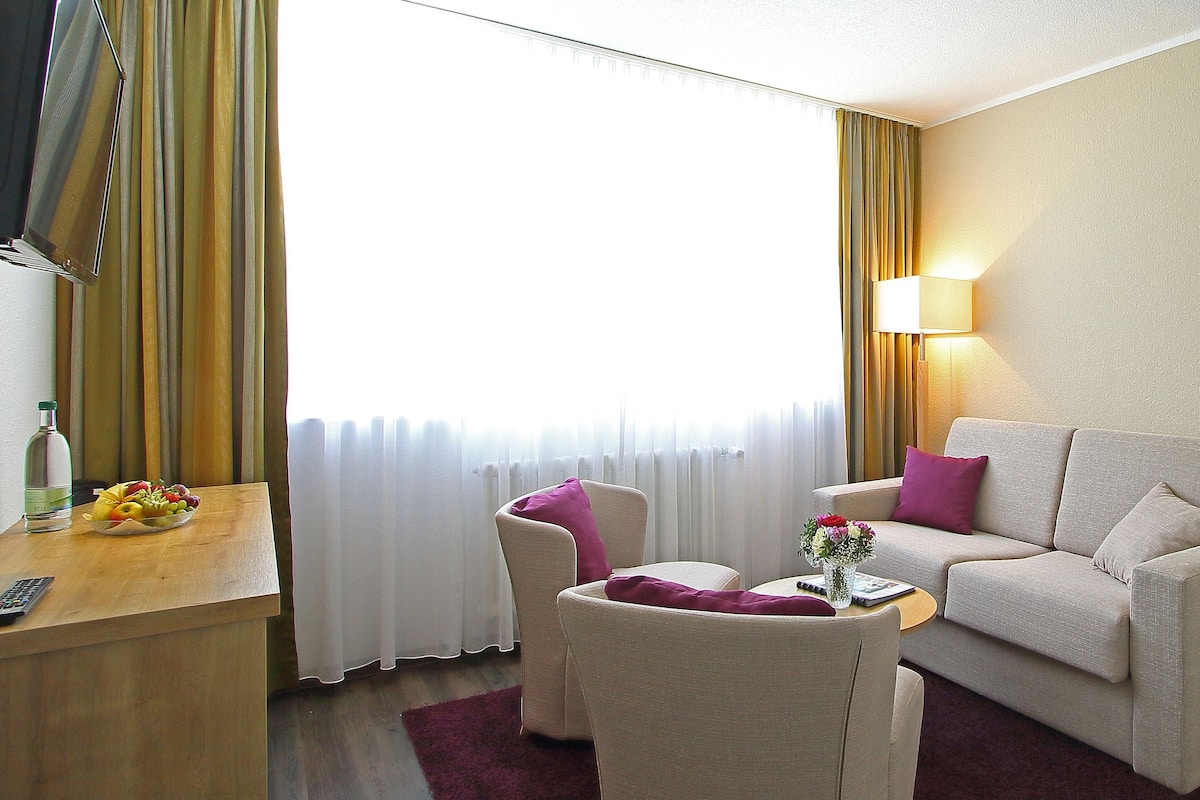 Une chambre d’hôtel avec un lit, un canapé, une chaise, une petite table ornée d’un vase de fleurs et une lampe.