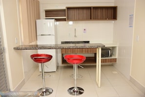 A compact kitchen with a white refrigerator, a microwave, and wooden cabinets. There are two red bar stools with metal bases. - Reserva Caldas Lacqua 3 (Caldas Novas)