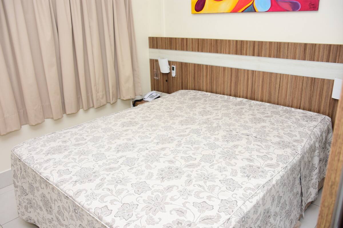 Un lit bien fait, recouvert d’une couvre-lit à motifs floraux, avec une tête de lit en bois, un téléphone sur la table de chevet et une applique murale.