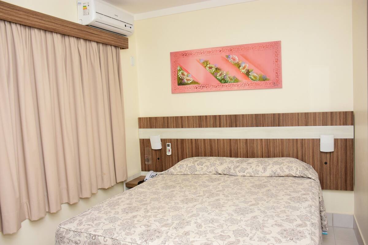 Une chambre d’hôtel avec un lit, des rideaux beiges, un climatiseur fixé au mur, une œuvre d’art à motifs floraux et une tête de lit en bois.