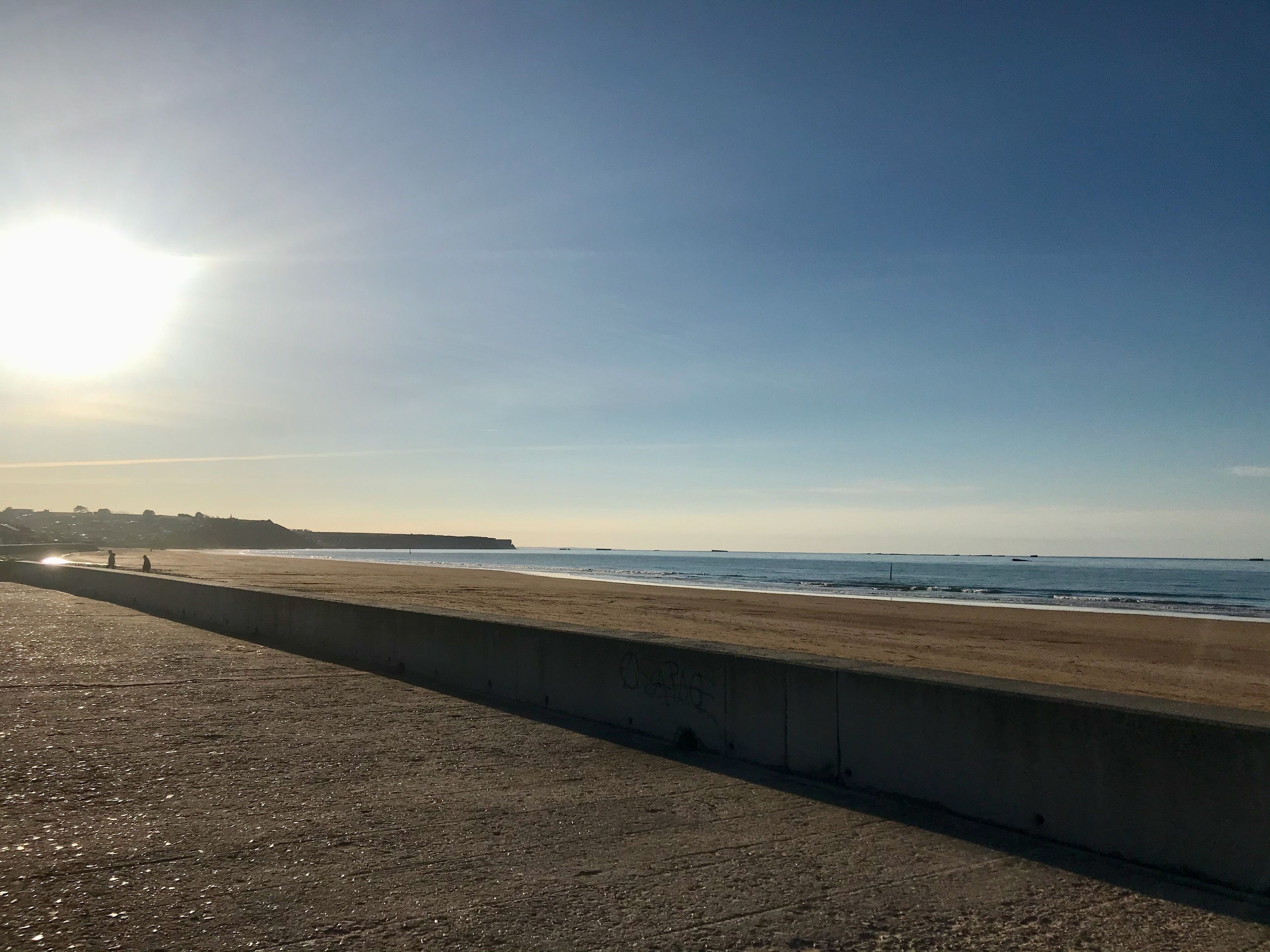 A beach with a concrete barrier, calm sea, and clear sky.