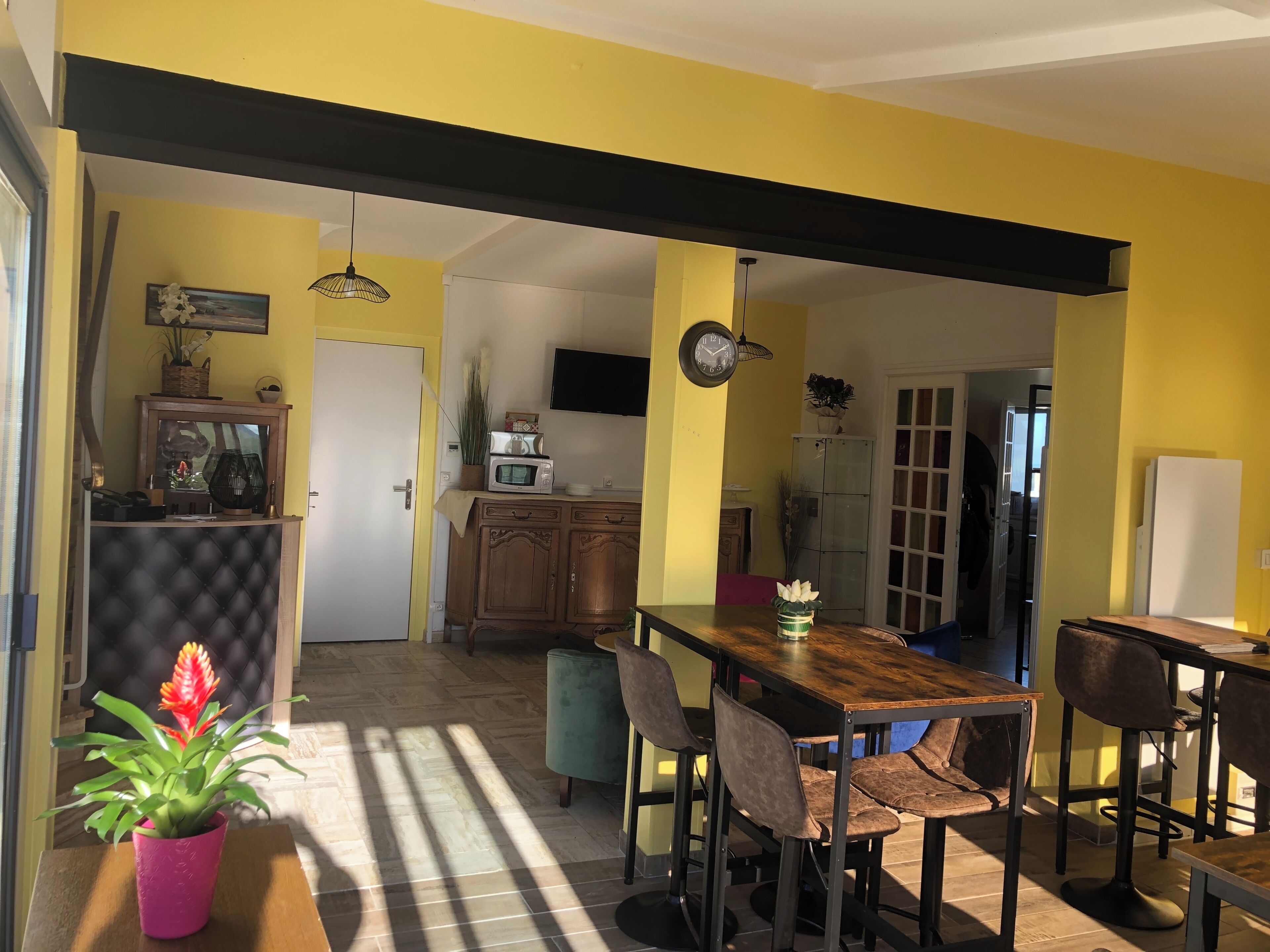 A dining area with a wooden table and chairs, a potted plant, and a kitchen in the background.