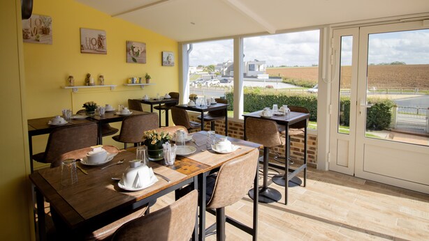Comedor con mesas dispuestas para una comida, pared amarilla con obras de arte y vistas al campo a través de amplias ventanas.