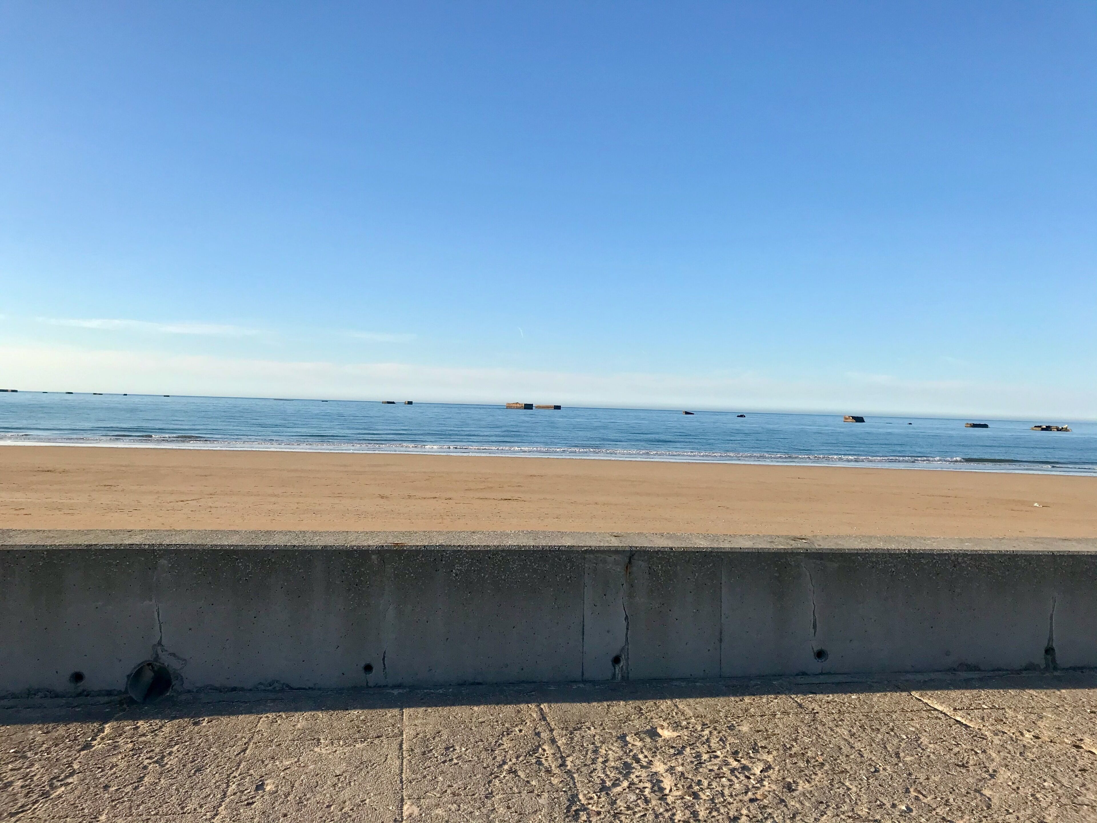 Een strand met een betonnen barrière op de voorgrond en een rustige zee op de achtergrond.