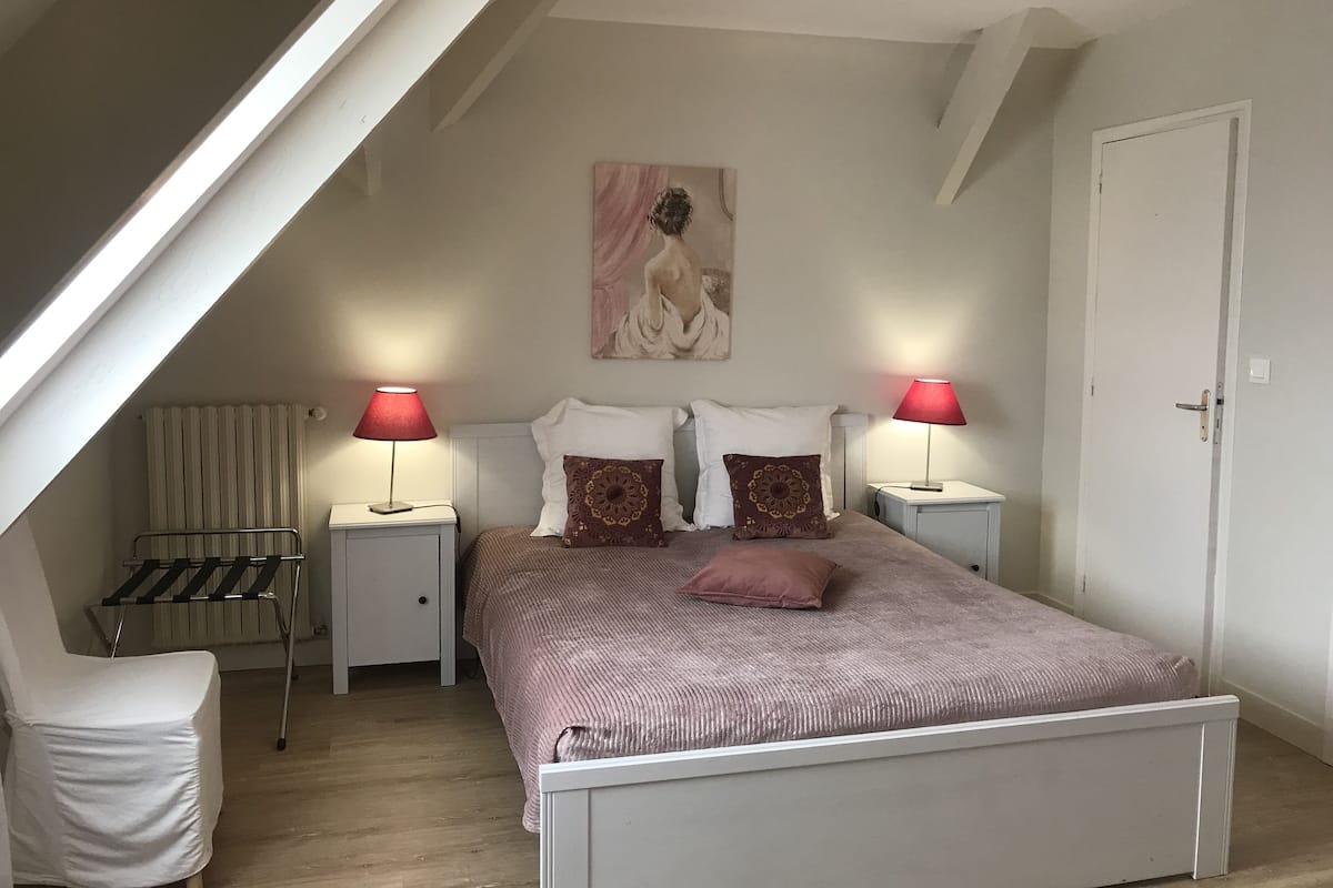 Un dormitorio con techo inclinado, una cama con colcha rosa, dos lámparas rojas, un radiador y un cuadro en la pared.