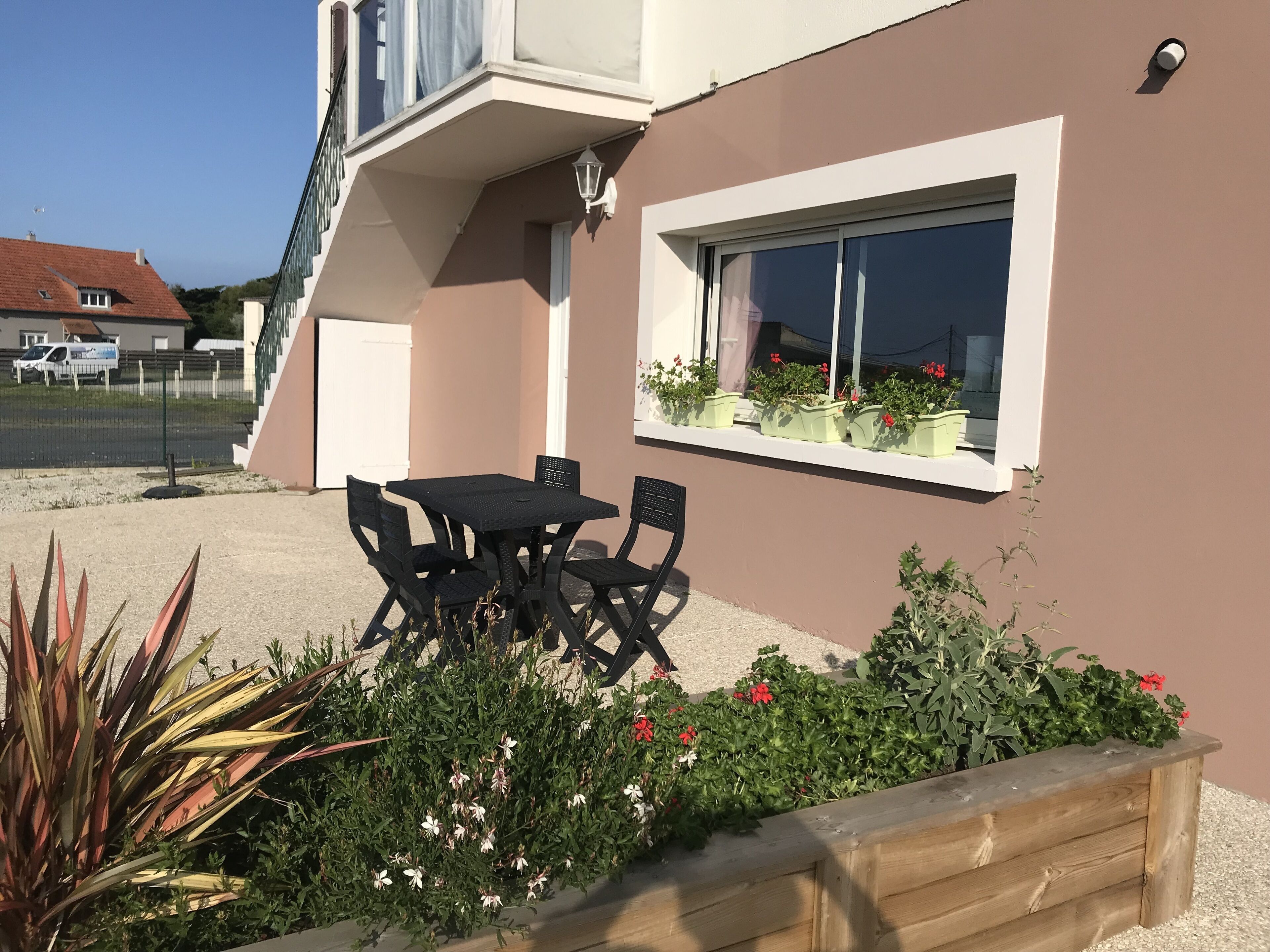 A patio with a table and chairs, a window with potted plants, and a garden with various plants and flowers.