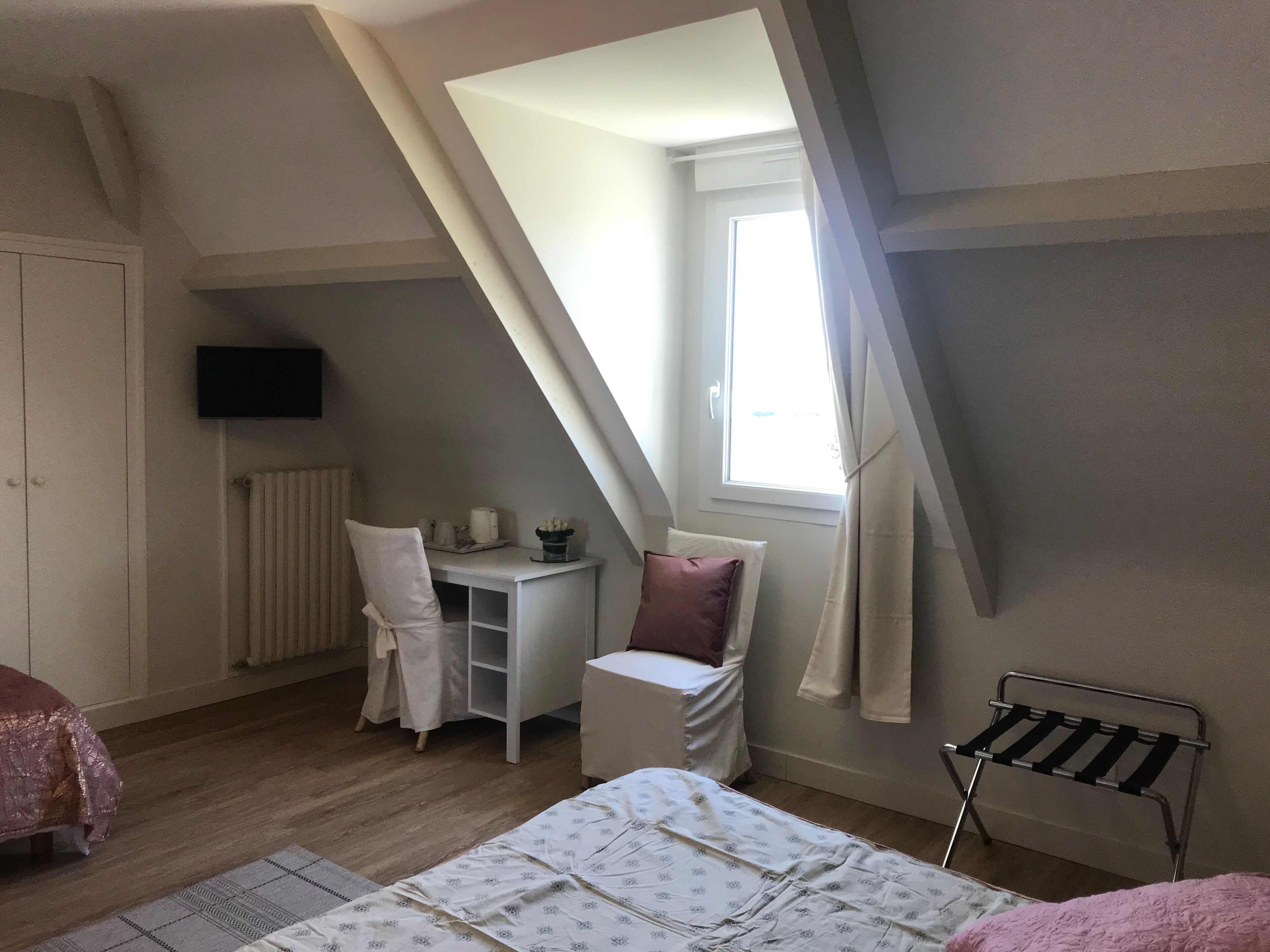 A bedroom with a sloped ceiling, a bed, a desk with a chair, a radiator, and a window with curtains.