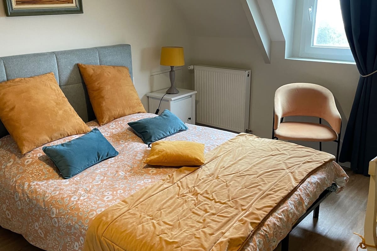 Un dormitorio con cama, almohadas, una mesita de noche, lámpara, silla, radiador y ventana con cortinas.