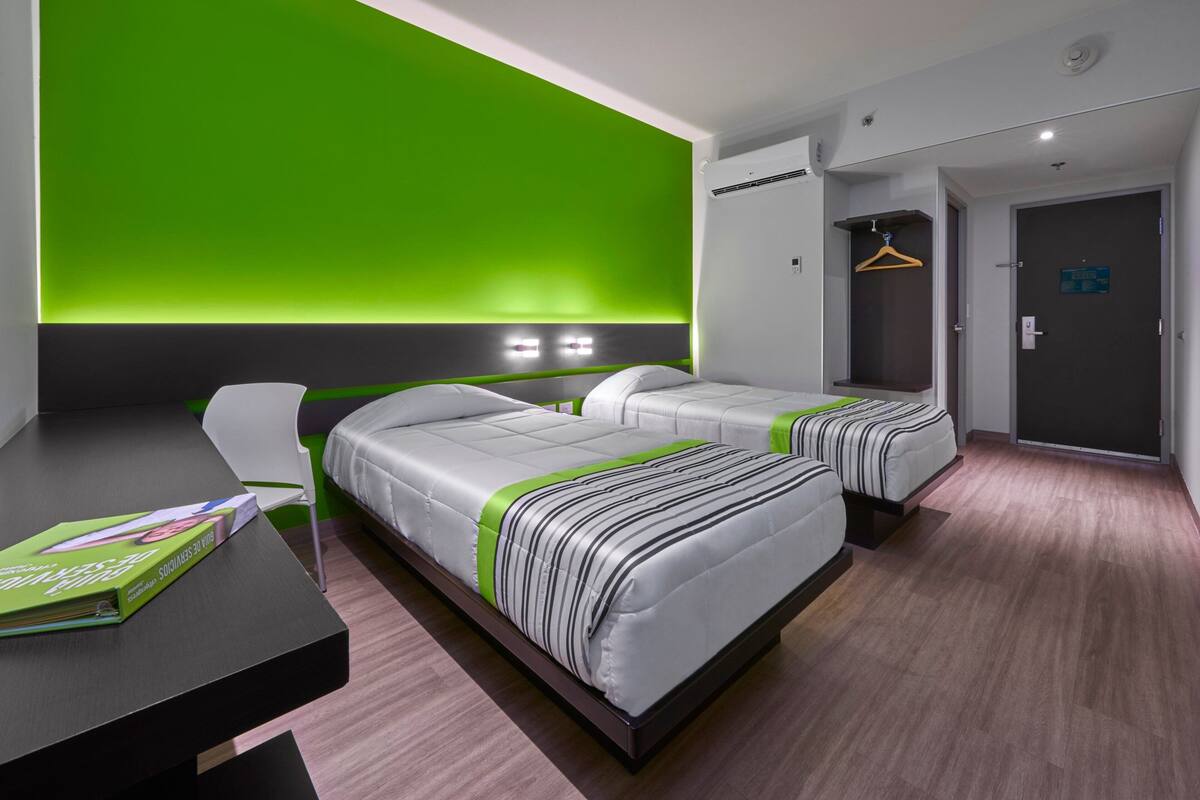 A modern hotel room with two beds, a desk, and a green accent wall.