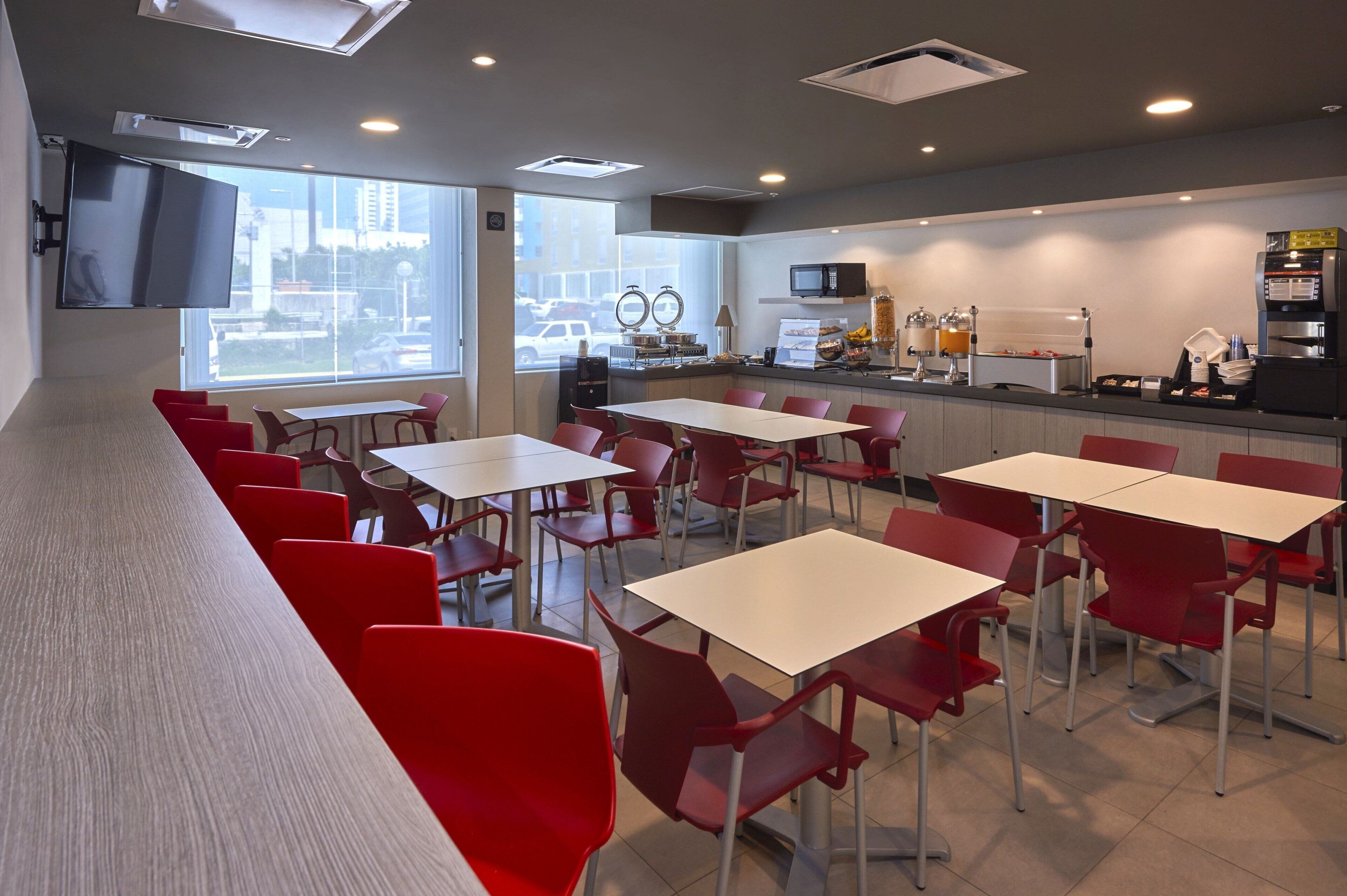 A modern dining area with red chairs and white tables, a flat-screen TV mounted on the wall, and a kitchen counter with a microwave and coffee machine.