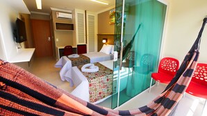 A hotel room with two beds, a glass partition, a red chair, a hammock, and a TV on the wall. - Pousada Recanto dos Corais (Ipojuca)
