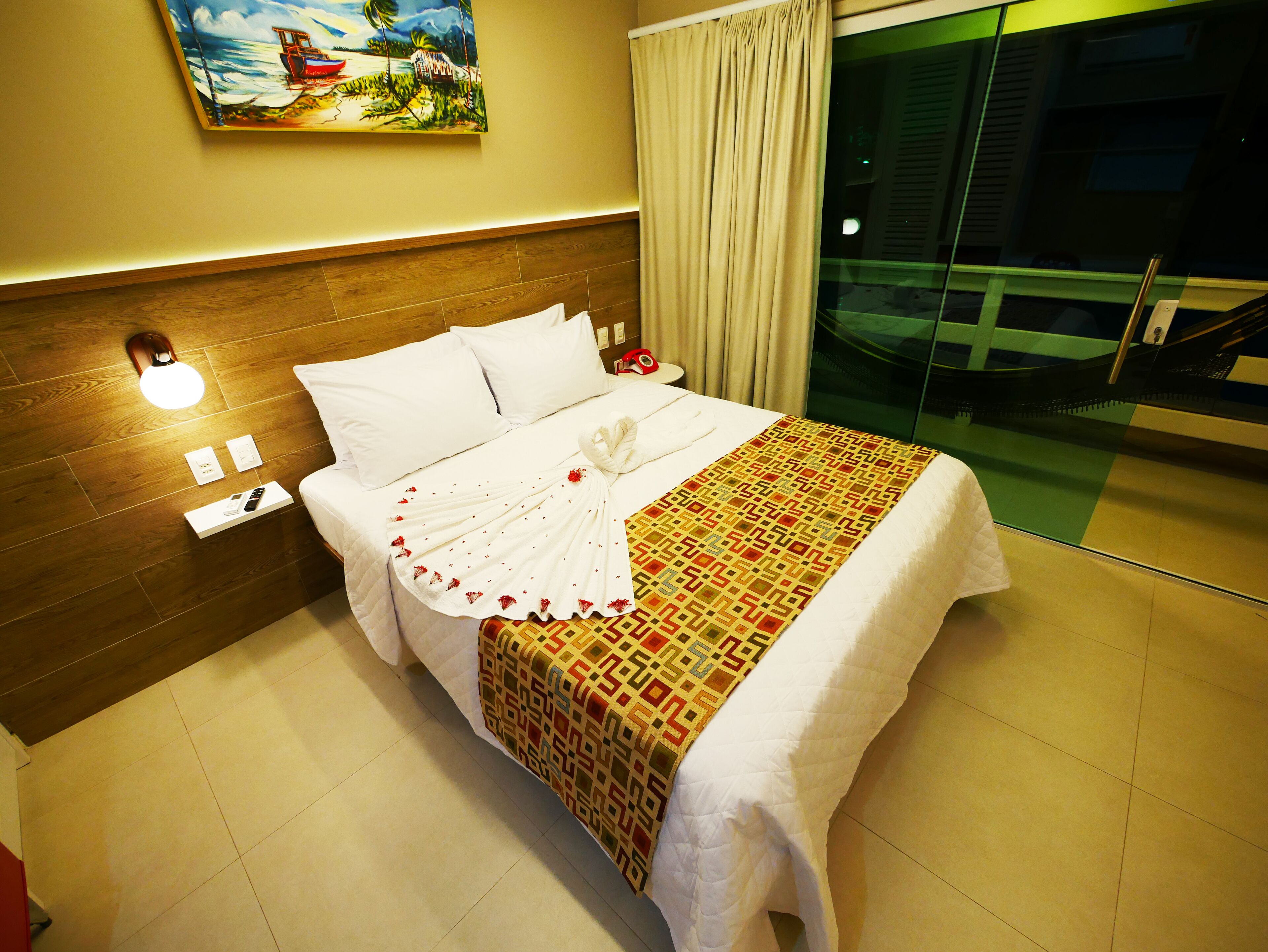 A neatly made bed with a patterned blanket, a white towel, and a remote control on the nightstand.