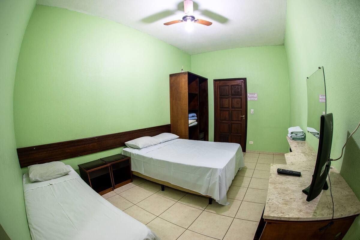 A small, well-lit room with a single bed, a wooden wardrobe, a desk with a mirror, and a ceiling fan.