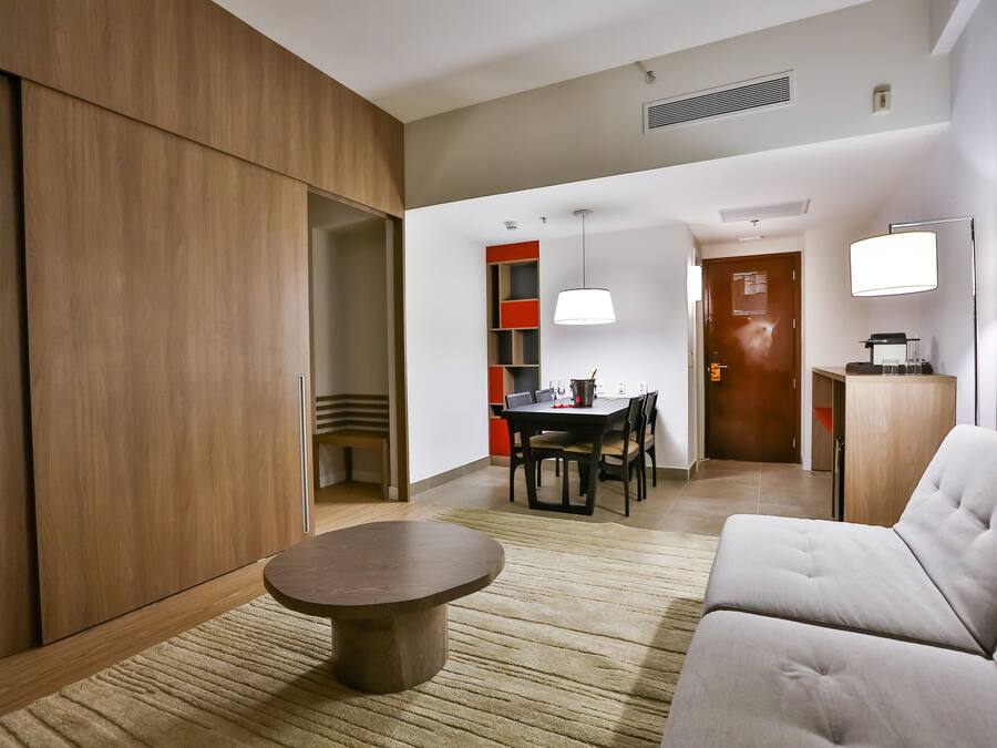 Una habitación de hotel moderna con un armario de madera, una mesa redonda, un sofá, un área de comedor con mesa y sillas, y un minibar.