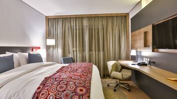 Una habitación de hotel con una cama grande, un escritorio, una silla y un ventanal con cortinas.